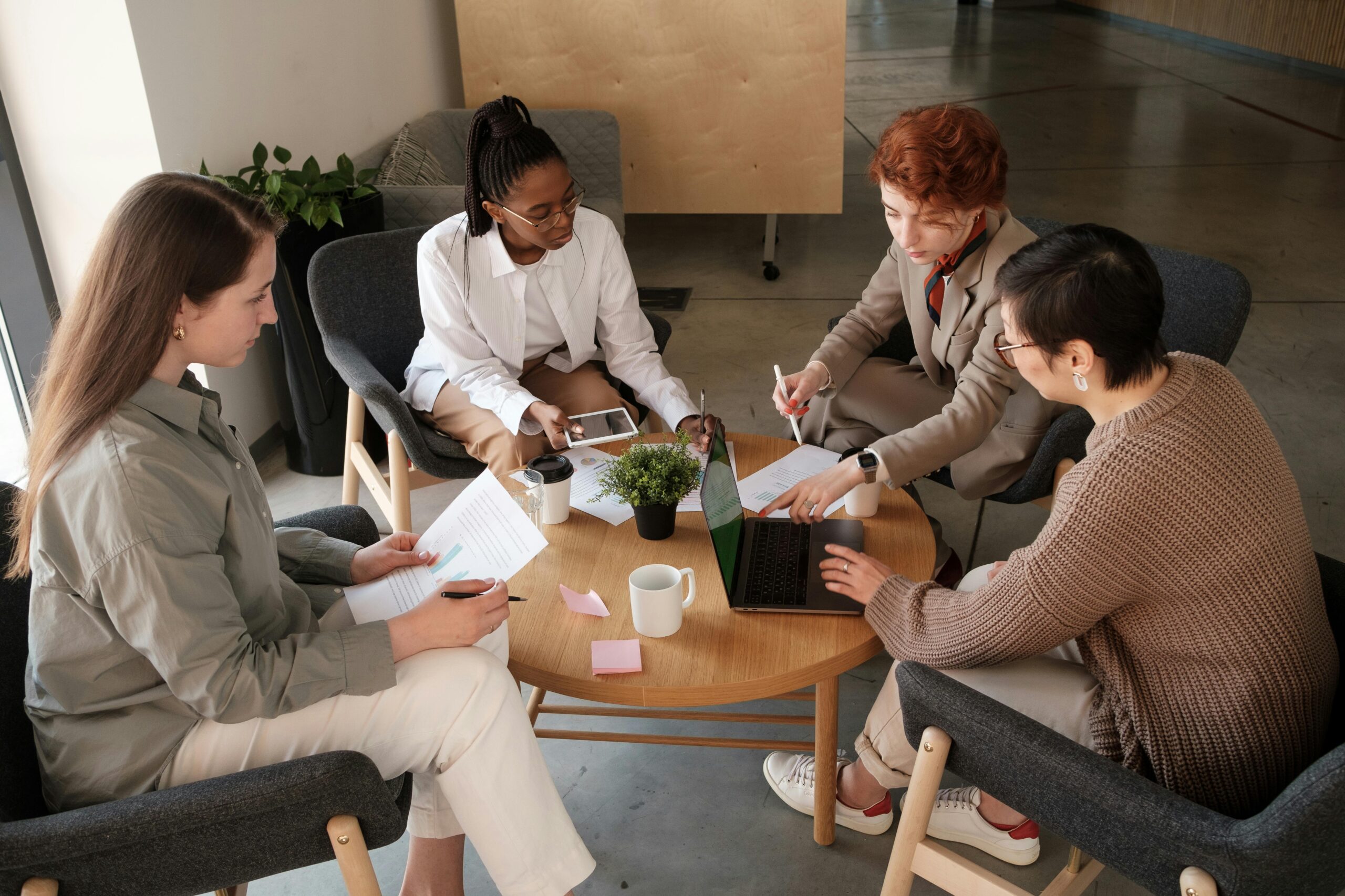 4 women in business attire sitting around table with coffee and laptops
