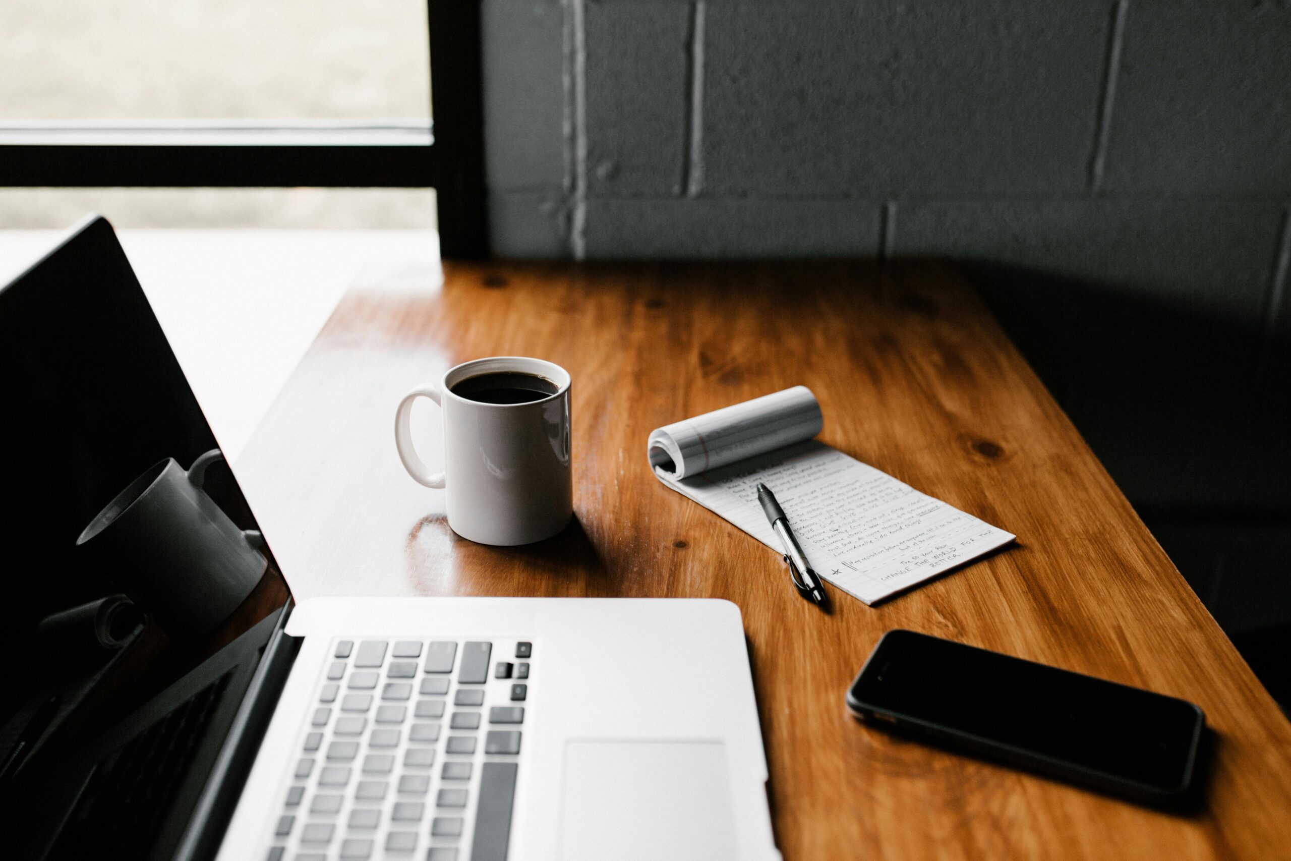 Laptop on wooden table next to coffee mug and notebook with pen