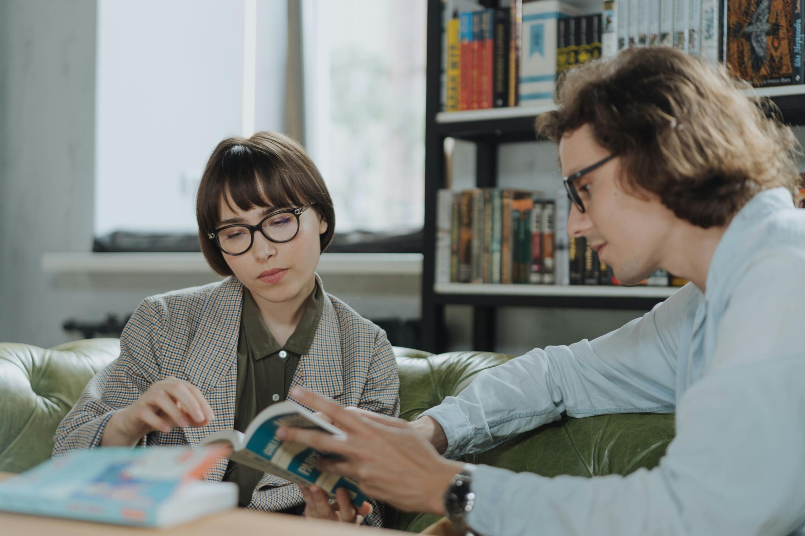 Two young adults sitting on a green couch in a library, discussing and reading a book together with shelves of books in the background.