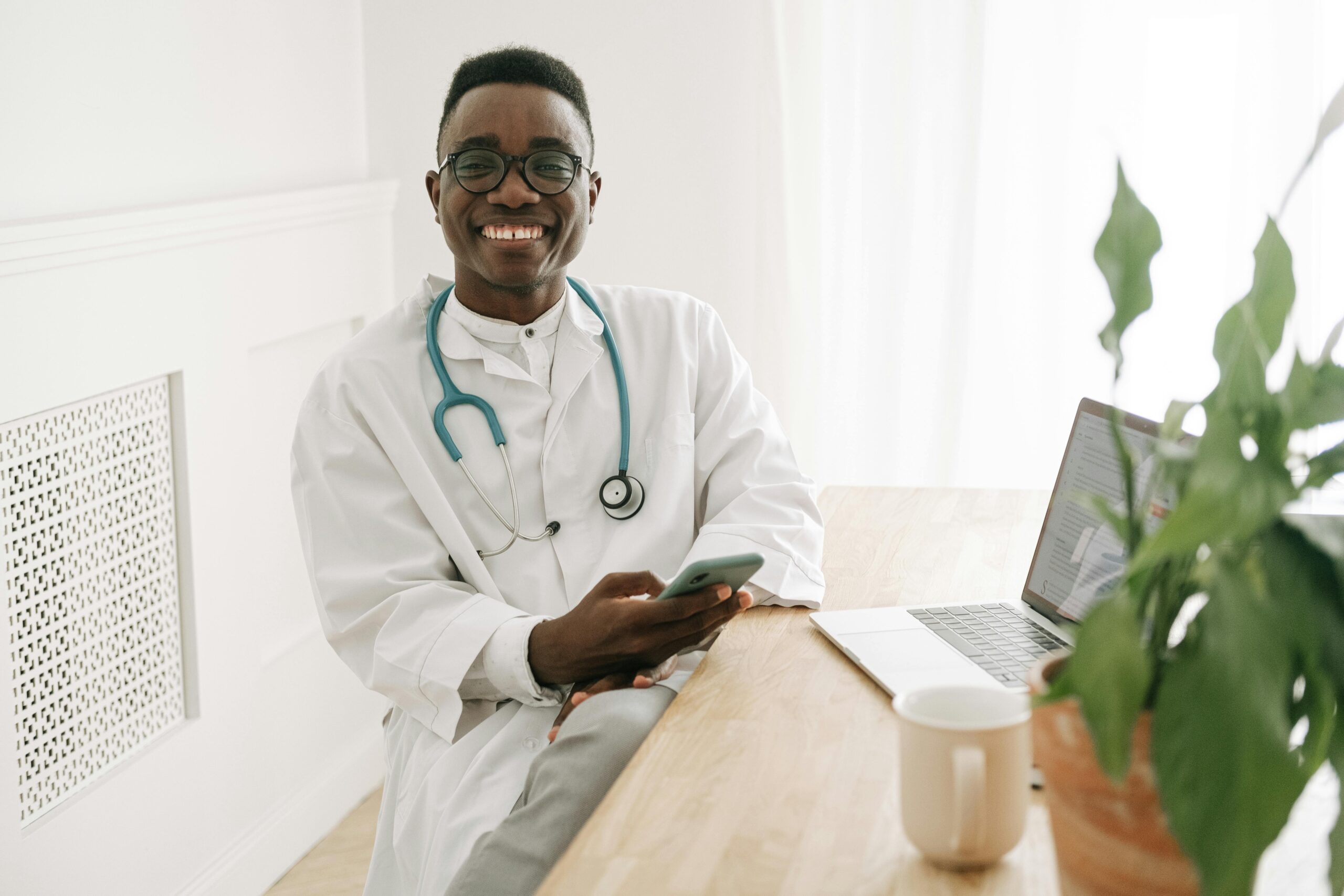 A young male doctor wearing a stethoscope around his neck smiles while looking at someone and holding a smartphone in his hand.