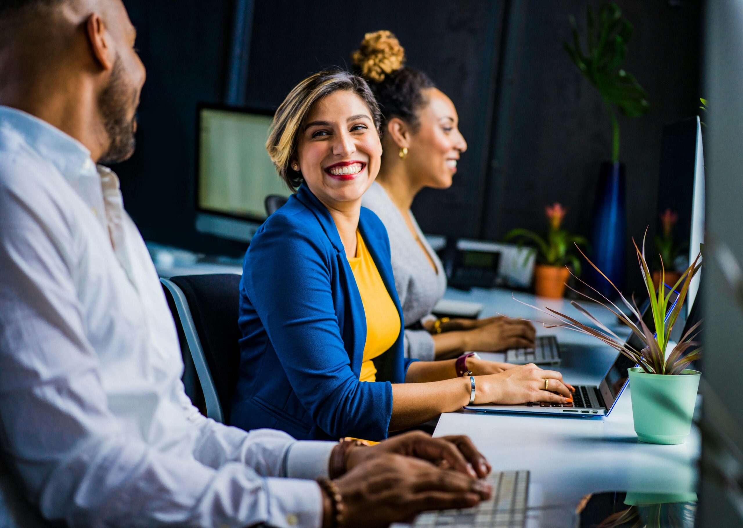 3 people dressed in business attire sitting in a line on laptops. One person is smiling direct to camera