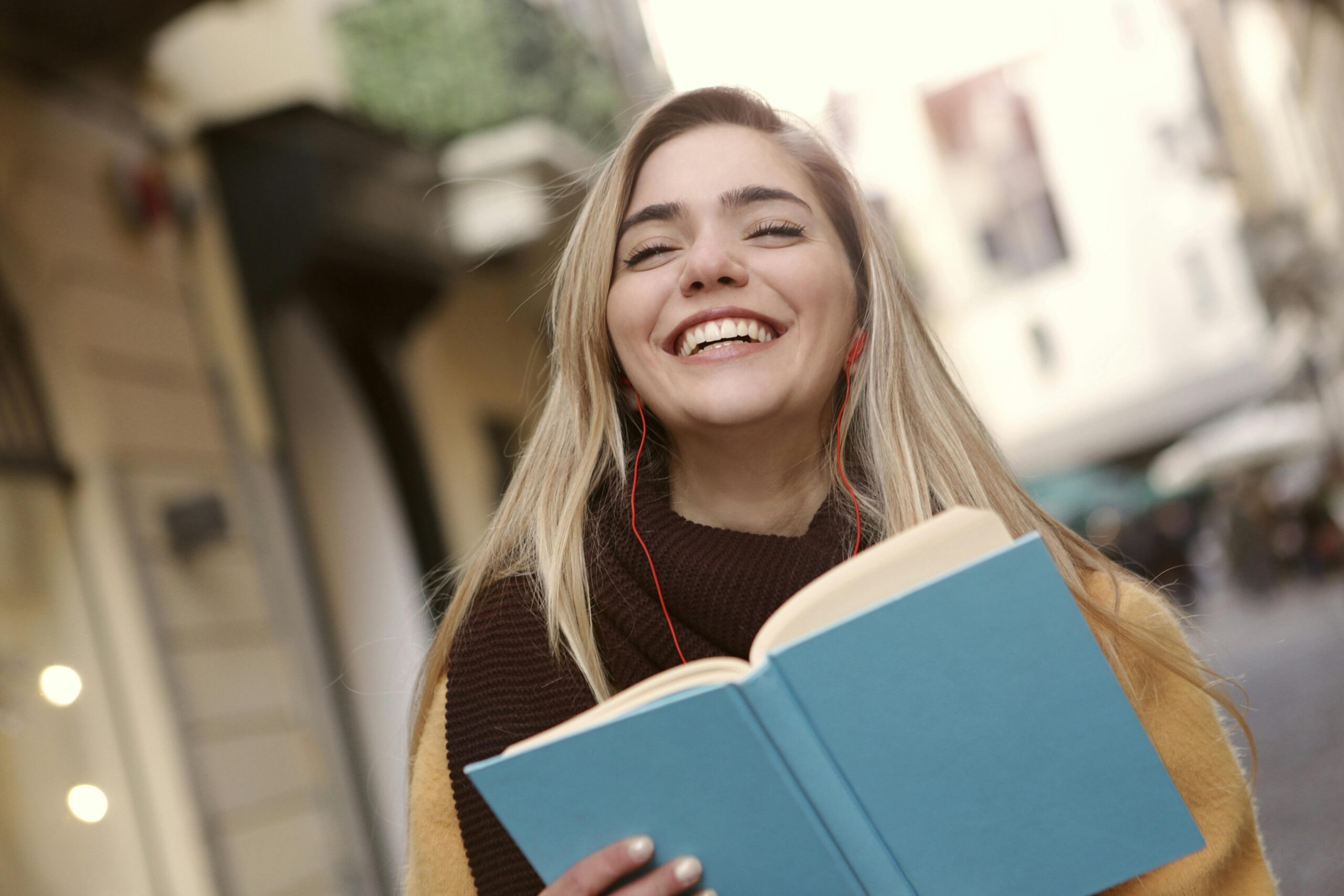 Smiling young woman with long blonde hair wearing earphones, holding an open blue book while standing outdoors in a city street.
