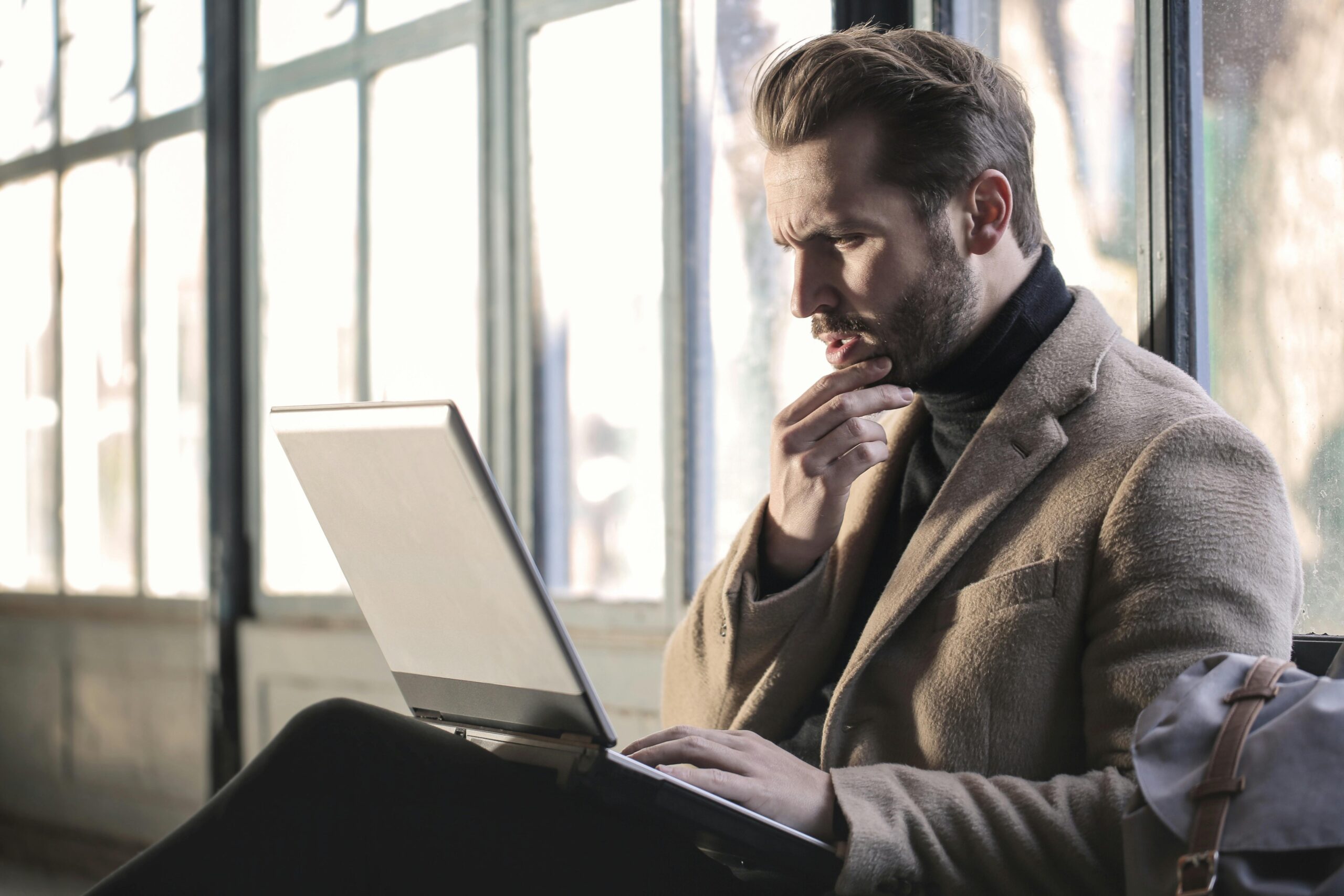 Man in business attire sitting next to window working on laptop