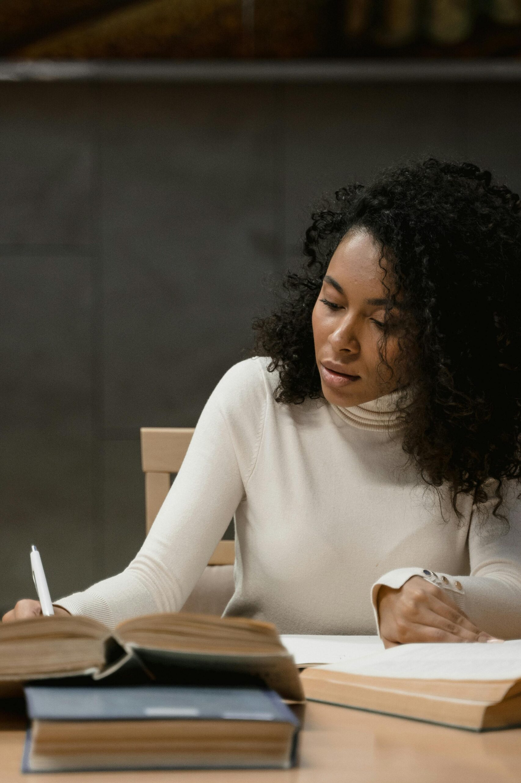 A young woman sitting at a study table, writing in a notebook surrounded by open books, focused on her study.