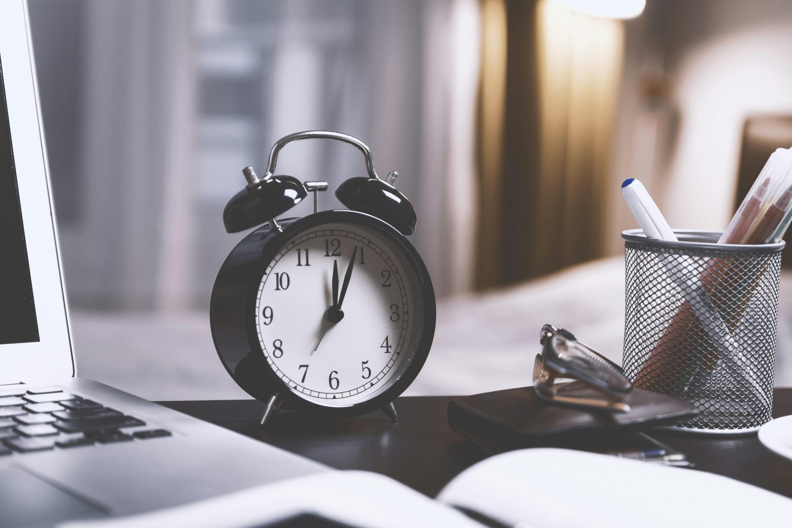 Classic analogue alarm clock sitting on office desk next to cup of pens and laptop