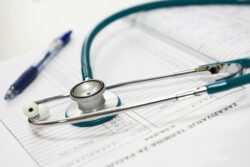 A stethoscope rests on a medical form with printed text. A blue pen lies nearby, suggesting a healthcare setting. The tone is clinical and professional.